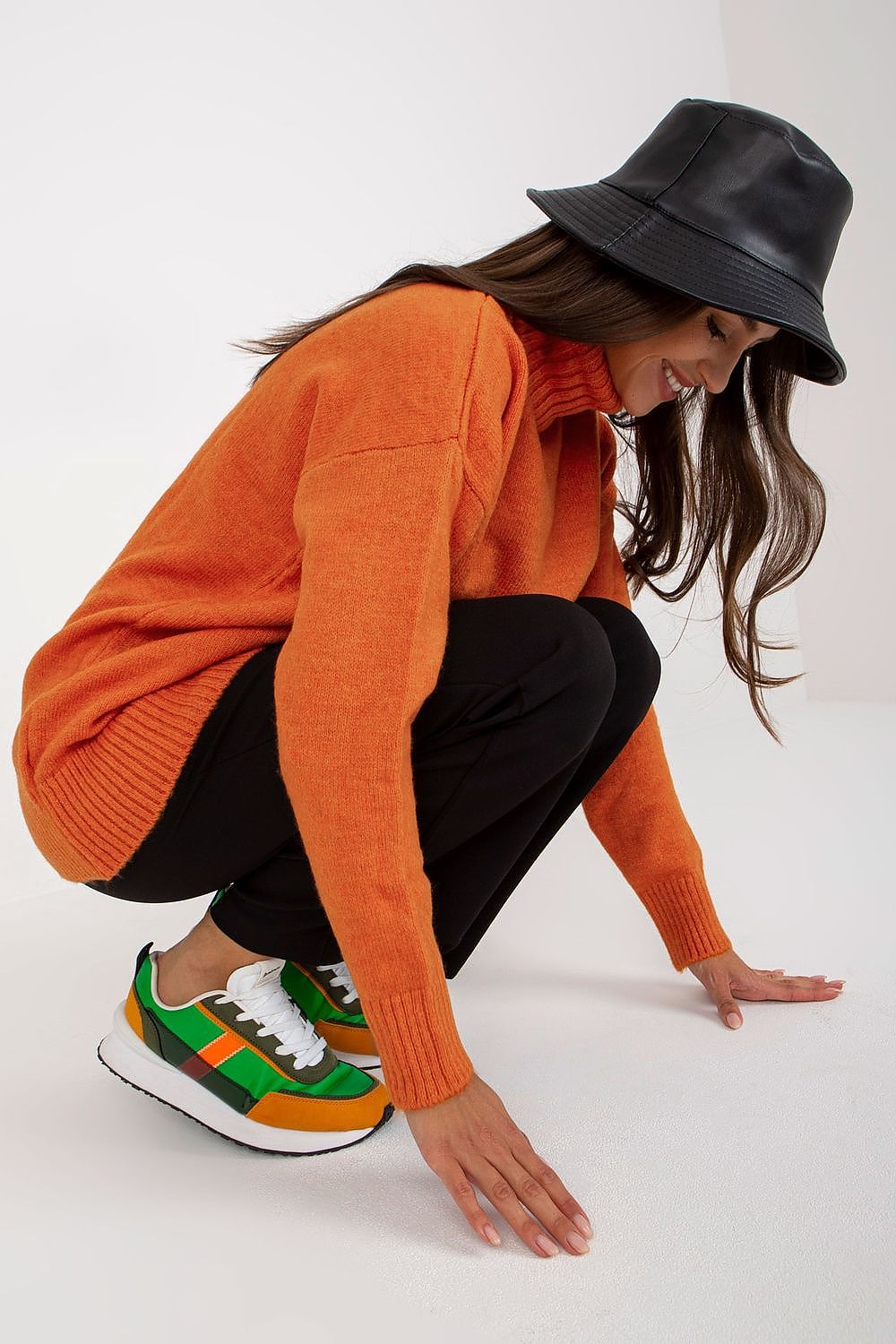 Model crouched gracefully in an elegant rust turtleneck sweater by Rue Paris, complemented by vibrant sneakers and a black hat, embodying a sophisticated autumn look.