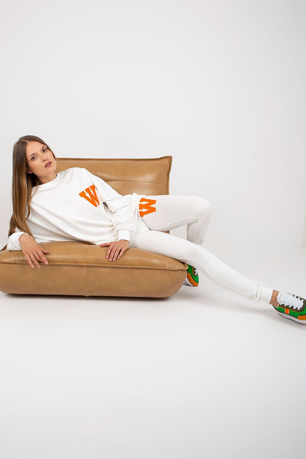Elegant model lounging in a stylish white sweatsuit set, adorned with vibrant orange 'W' patches, showcasing a luxurious blend of comfort and chic fashion.