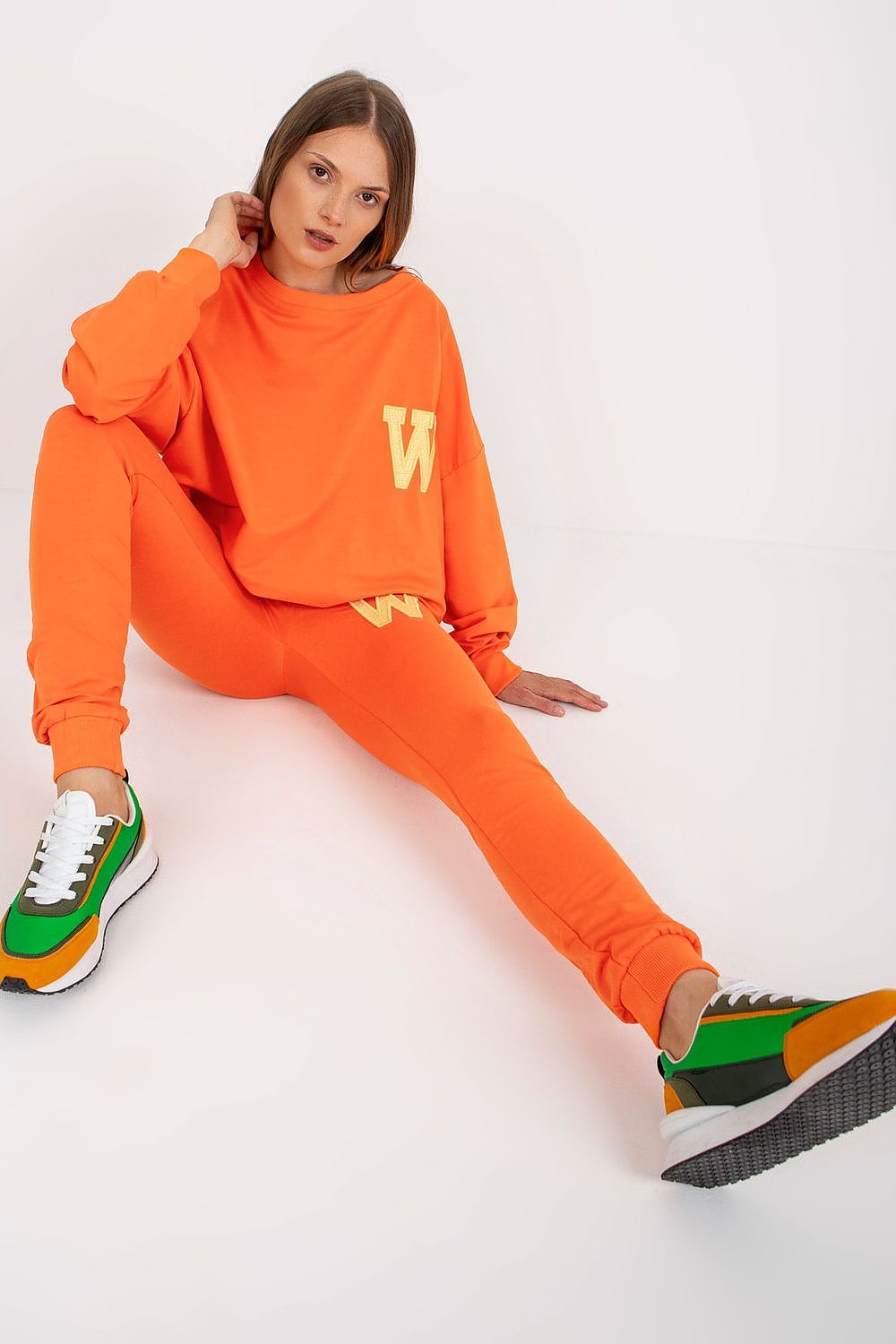 Elegant model showcasing an orange cotton-spandex sweatsuit set featuring a loose-cut sweatshirt and stylish long pants, complemented by trendy sneakers.