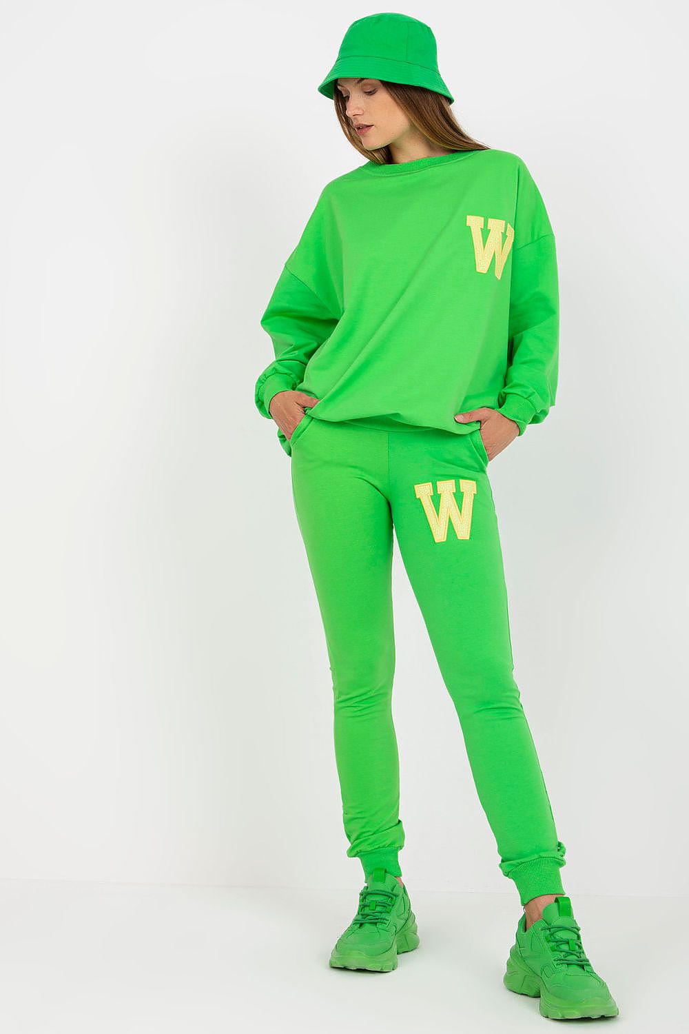 Chic model in a vibrant green cotton-spandex sweatsuit set, featuring a loose-cut sweatshirt with a patch and matching long pants, adorned with a fashionable bucket hat.
