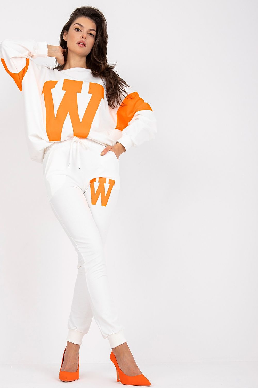 Luxury women's tracksuit set featuring a loose cut sweatshirt with bold orange 'W' logo and tailored white pants, styled with vibrant orange heels against a clean white background.