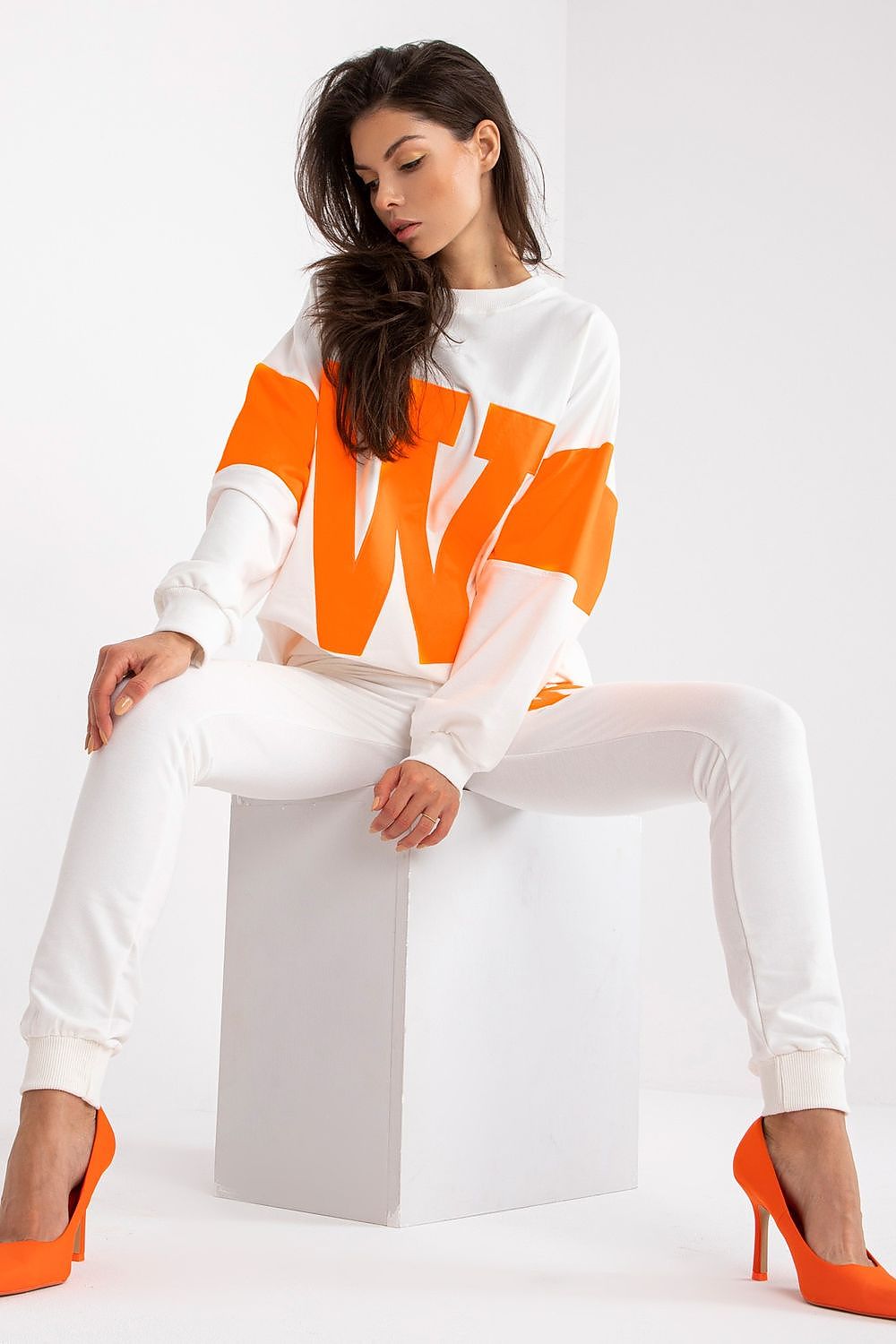 Chic women's tracksuit set in a relaxed fit, showcasing an oversized sweatshirt with orange accents and fitted white trousers, complemented by stylish orange heels while seated in a minimalist setting.