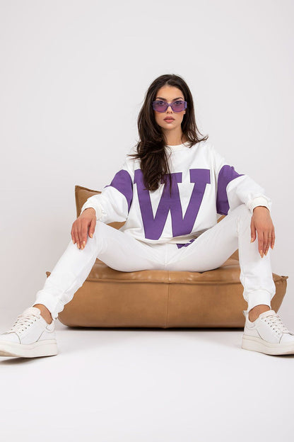 Luxury women's tracksuit set featuring a loose cut sweatshirt in white with bold purple lettering, paired with tailored white pants, styled with trendy sneakers.