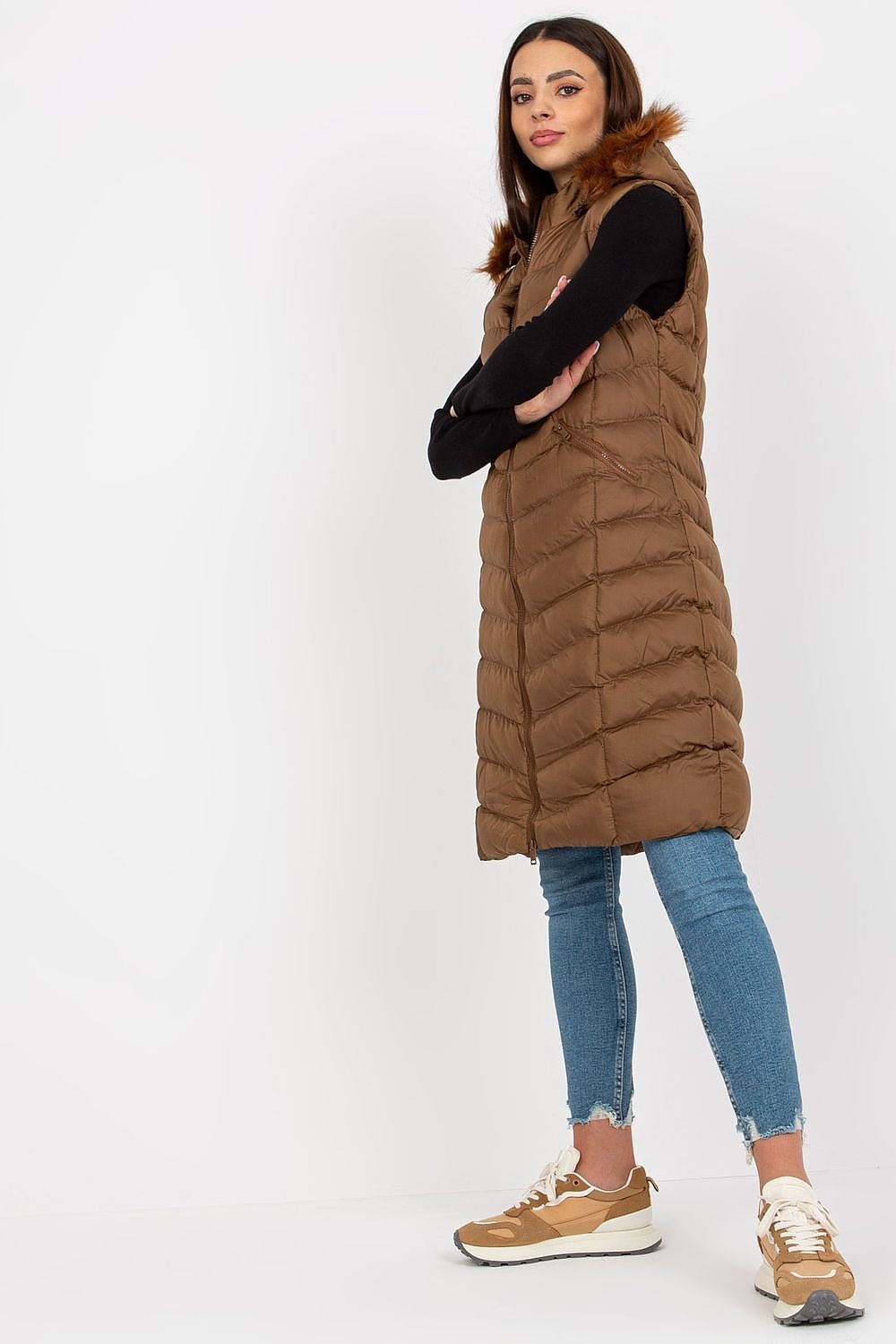 Elegant model showcasing the Luxury Rue Paris Down Long Zippered Vest in a rich brown hue, featuring a detachable hood and stylish slide pockets, paired with chic denim and fashionable sneakers.