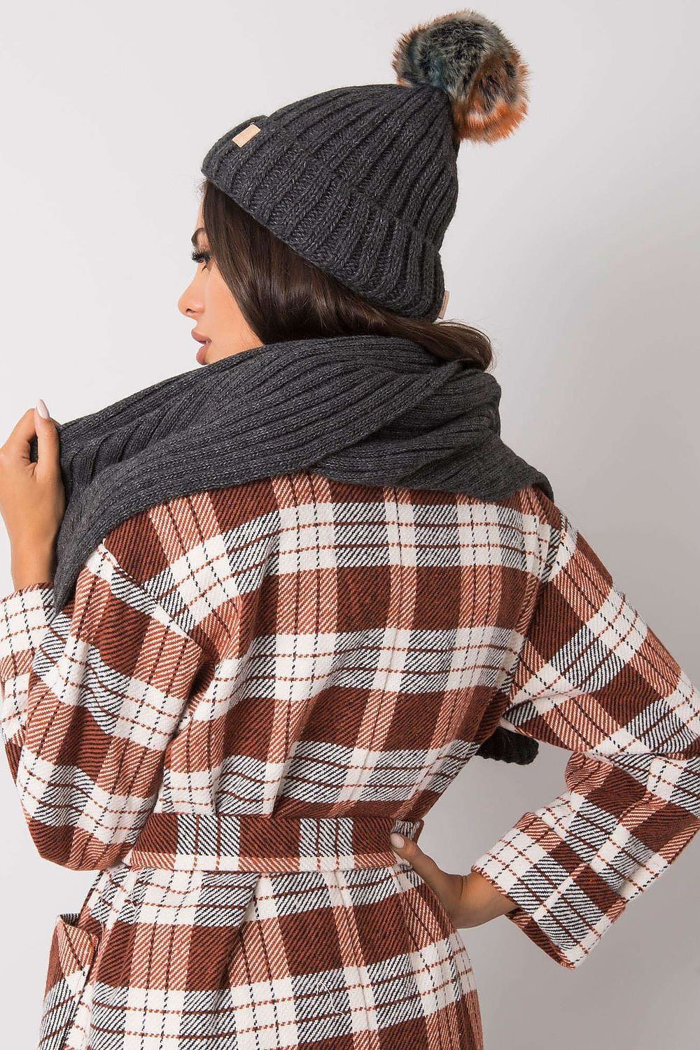 Elegant model showcasing a luxurious winter hat and scarf set in dark grey, featuring a textured knit pattern and a chic pom-pom detail, perfectly styled with a fashionable checked coat.