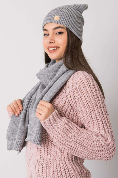 Elegant women's winter set featuring a luxurious grey beanie and scarf made from 100% acrylic. Model showcases the stylish accessories paired with a pink knitted sweater, highlighting the sophisticated design perfect for winter fashion.