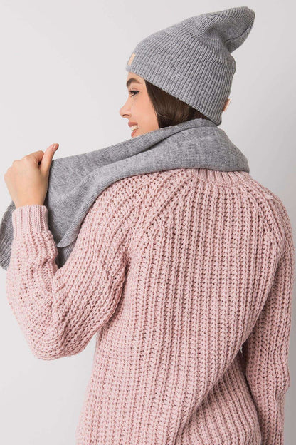 Chic view of a women's winter hat and scarf set in grey, made of 100% acrylic. The model displays the accessories gracefully draped over her shoulders, complemented by a soft pink sweater, embodying modern winter elegance.