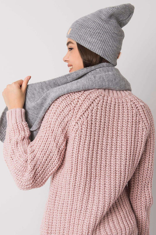 Chic view of a women's winter hat and scarf set in grey, made of 100% acrylic. The model displays the accessories gracefully draped over her shoulders, complemented by a soft pink sweater, embodying modern winter elegance.