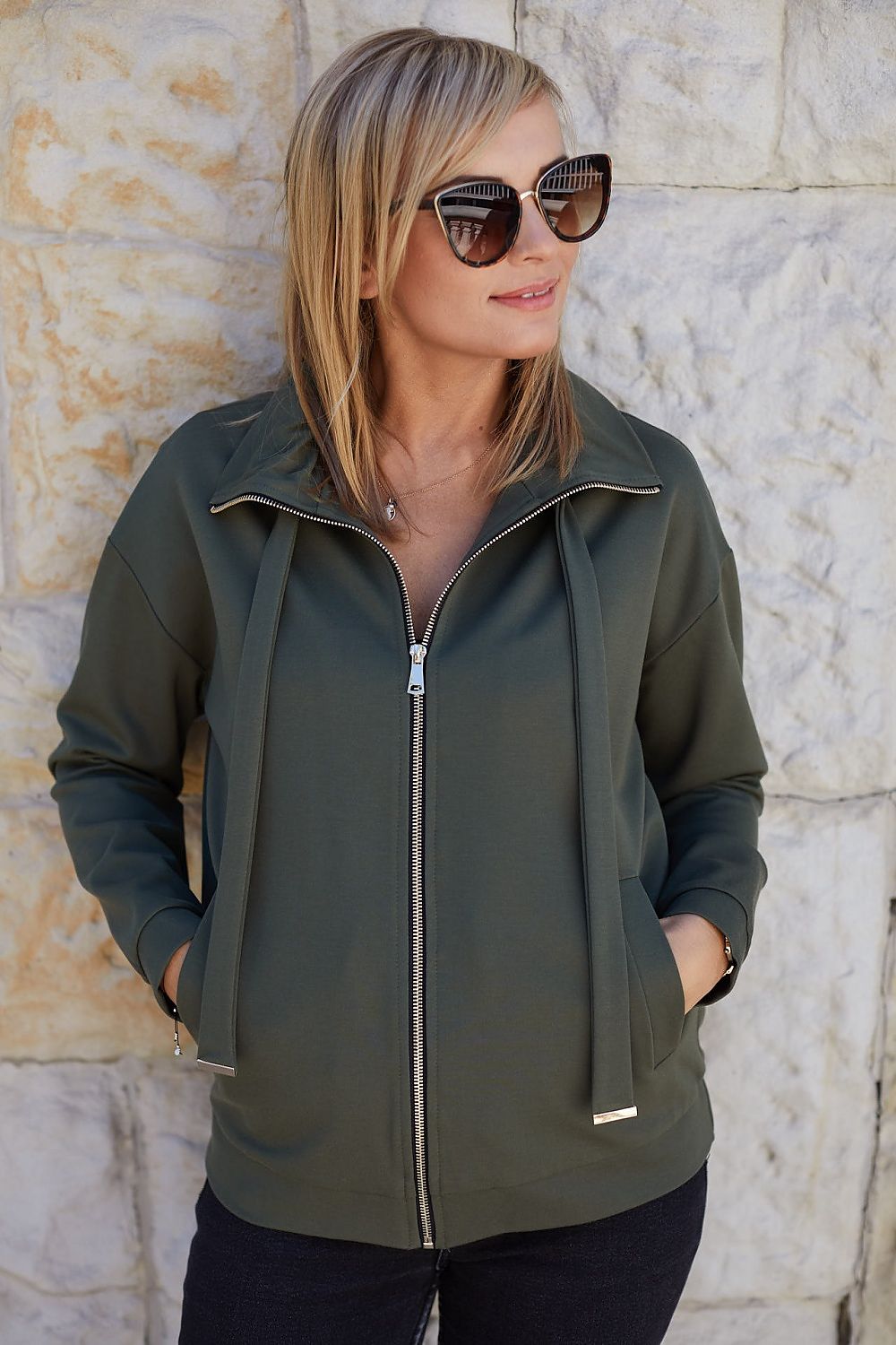Stylish woman wearing a La Aurora Luxury Unbuttoned Sweatshirt with metal hardware and turtleneck, posed against a stone wall, exuding sophistication with oversized sunglasses.