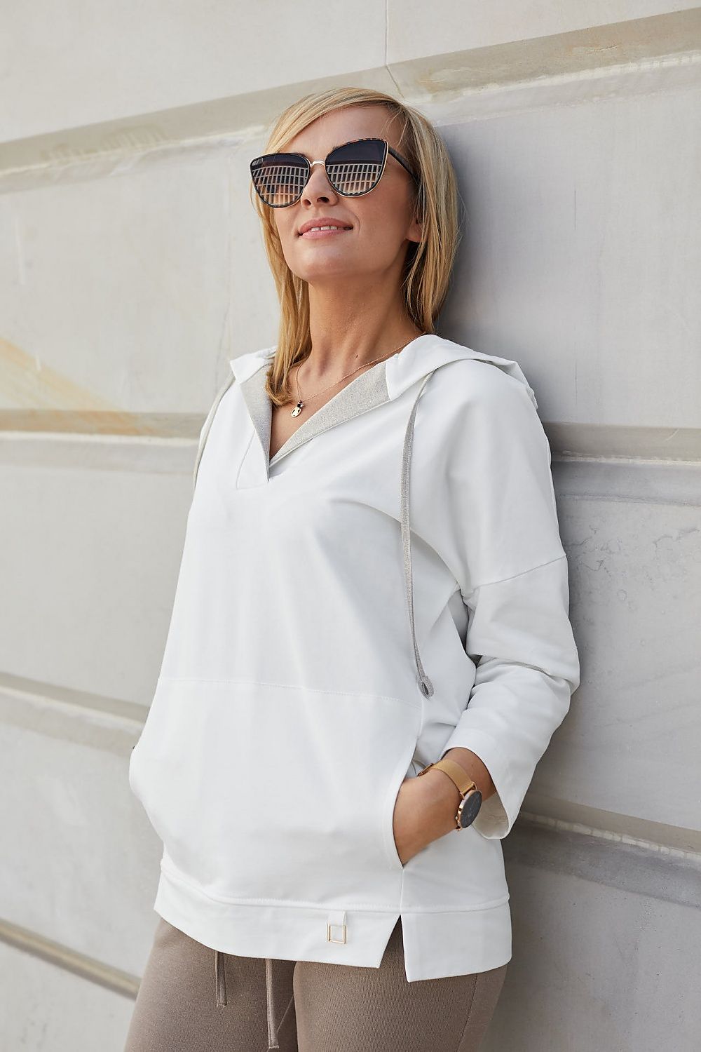 Elegant woman wearing a luxury beige hoodie sweatshirt with gold thread, showcasing a sporty-chic look and kimono sleeves, styled with fashionable sunglasses against a minimalist background.