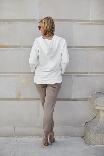 Back view of a fashionable woman in an exquisite beige hoodie, complemented by chic trousers and elegant footwear, radiating sophistication as she poses in front of a plain backdrop.