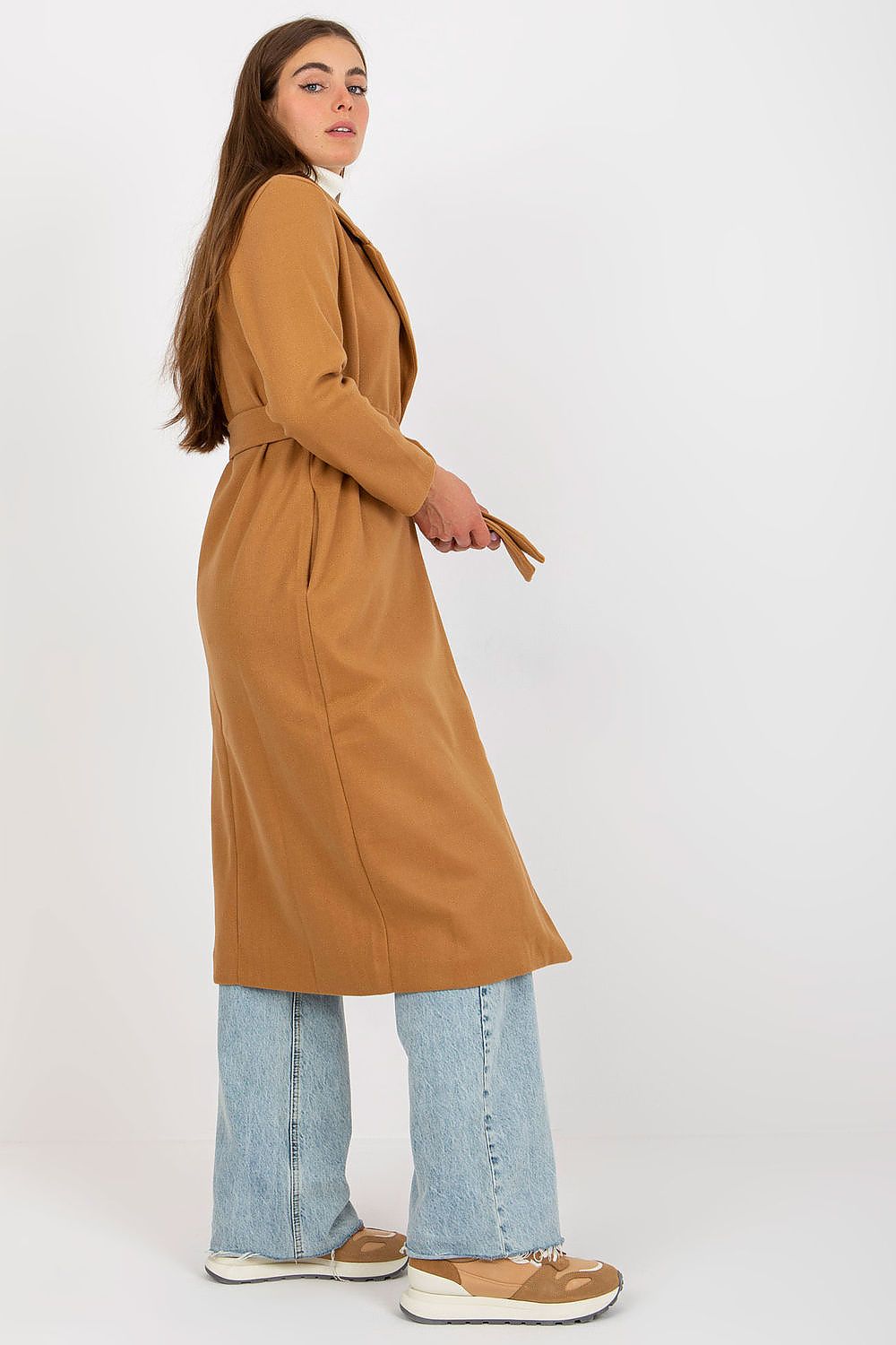 Side view of a stylish woman in an ochre coat featuring long sleeves and a tied waist, complemented by a white turtleneck and flared light blue jeans, embodying contemporary elegance.