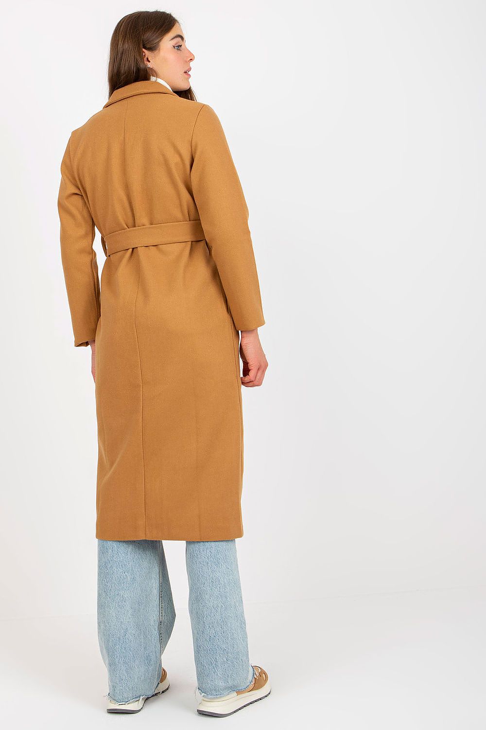 Rear view of a fashionable woman in an ochre long coat with a belt, creating a flattering silhouette, styled with a white turtleneck and relaxed fit light blue jeans, exuding luxury style.