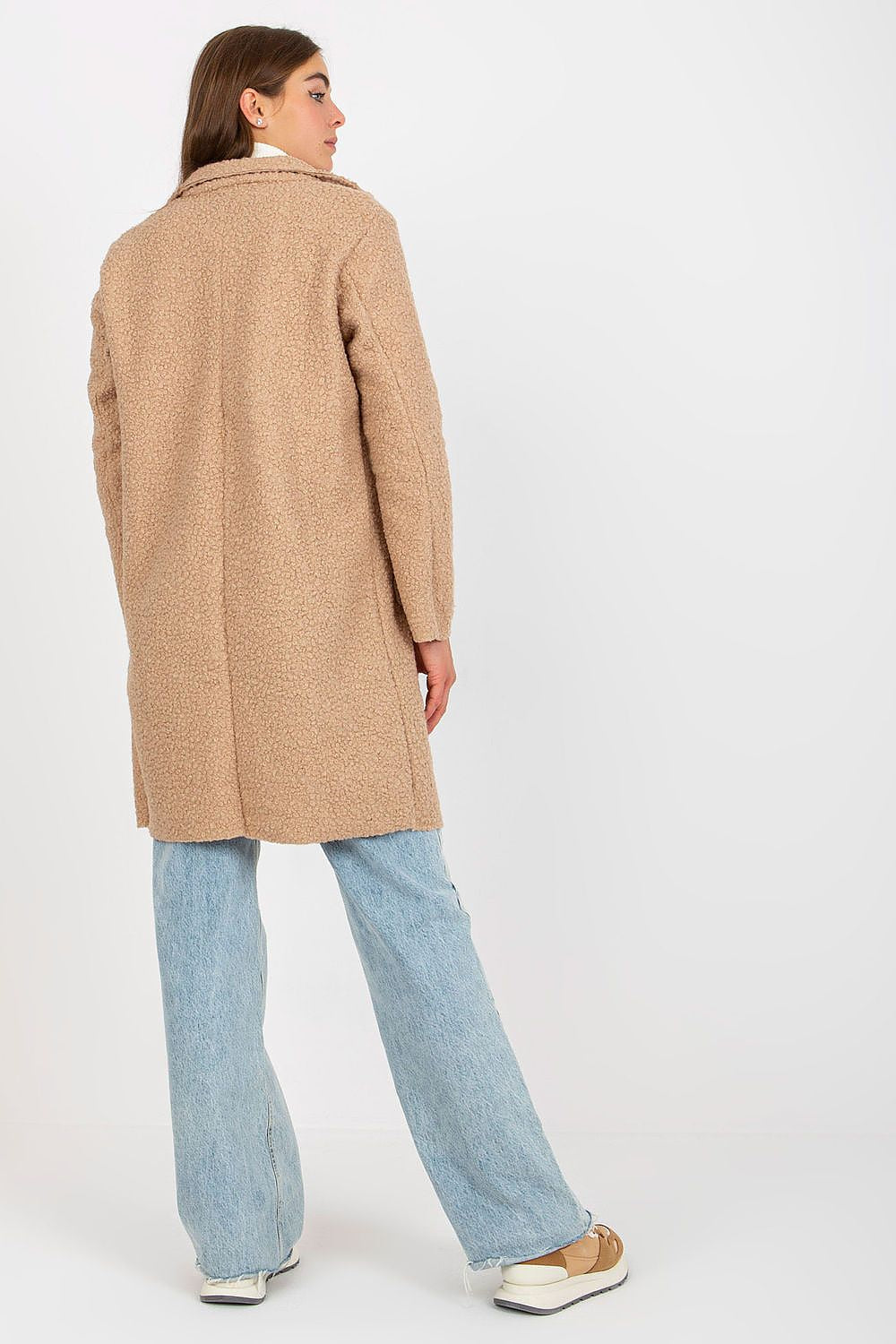 Elegant woman wearing an Och Bella luxury unlined buttoned coat in a soft beige hue, styled with long sleeves and patch slip pockets, paired with light denim pants and trendy sneakers, exuding a chic and sophisticated look.