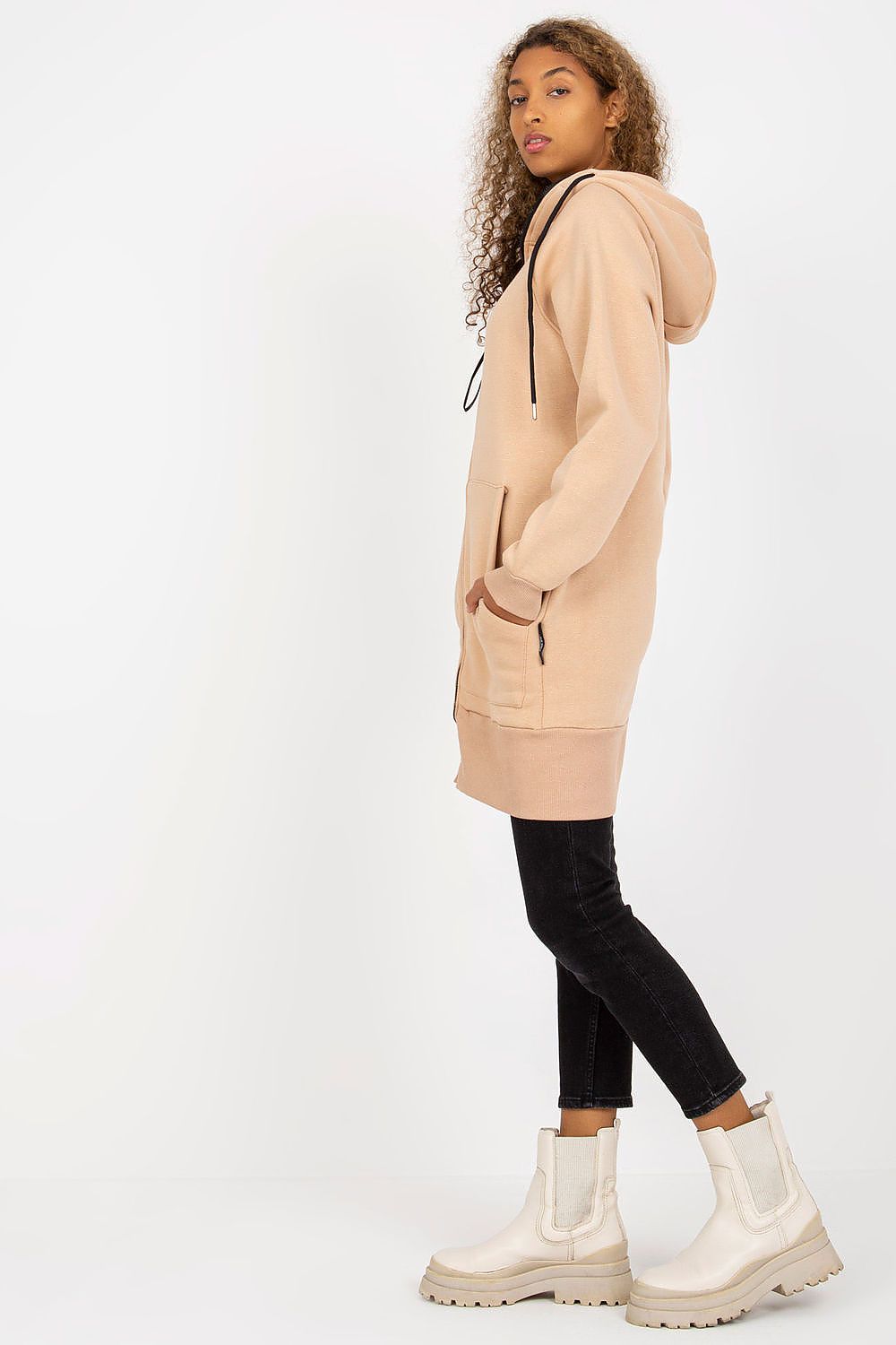 Model showcasing a luxury women’s hooded sweatshirt by Rue Paris in a chic beige shade, featuring a stylish extended cut and zippered front, paired with black skinny jeans and stylish ankle boots.