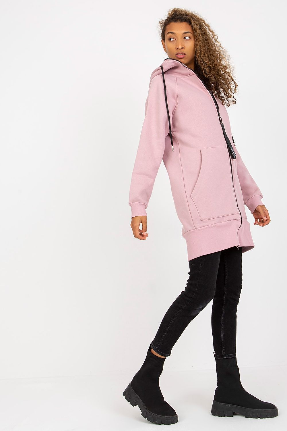 Model wearing a luxury women’s hooded sweatshirt by Rue Paris in soft pink, embellished with a zippered closure and side pockets, paired elegantly with fitted black jeans and chic ankle boots.