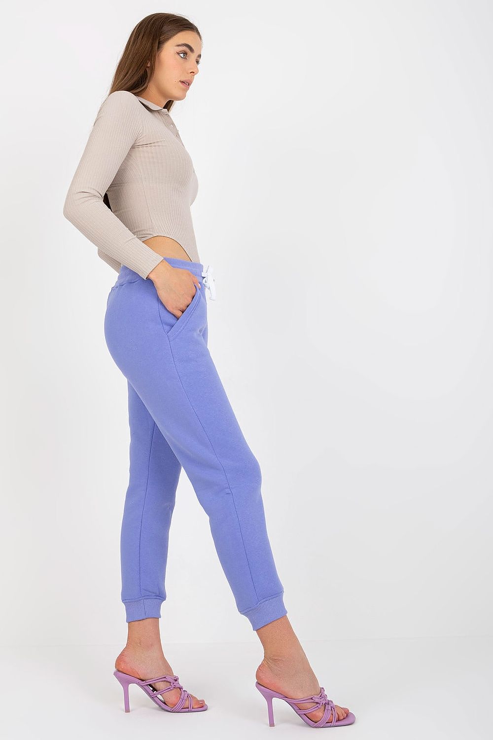 Side view of a sophisticated model wearing lavender cotton-spandex tracksuit trousers with waist tie and slip pockets, paired elegantly with a fitted ribbed top and stylish footwear.