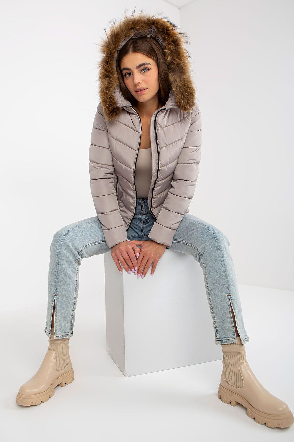 Chic model sitting confidently in a luxe polyester jacket with a faux fur trim, coupled with trendy denim, ideal for winter styling.