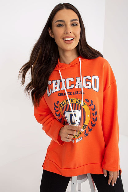 Elegant women's orange hoodie sweatshirt with a college logo print, designed in an asymmetrical style, paired with black skinnies and looks effortlessly trendy with a radiant smile.
