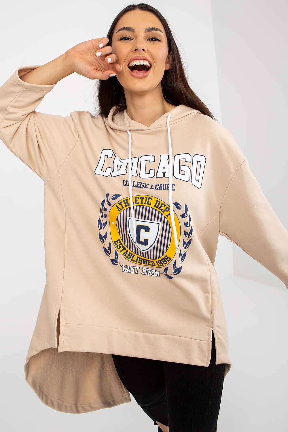 Cheerful woman in a luxurious beige hoodie sweatshirt with a college graphic, featuring an asymmetrical hem, styled with black leggings and exuding a casual chic aesthetic.