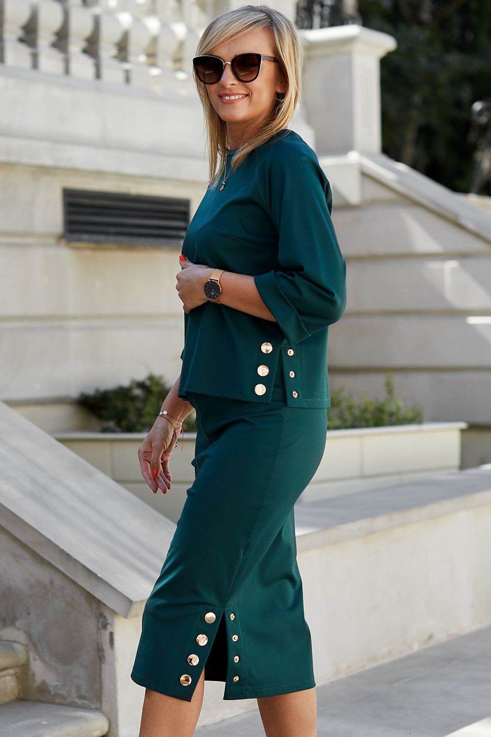 Side profile of a fashionable woman in a soft Morgan knit two-piece set featuring metallic press studs, smiling with confidence against a sophisticated architectural backdrop.