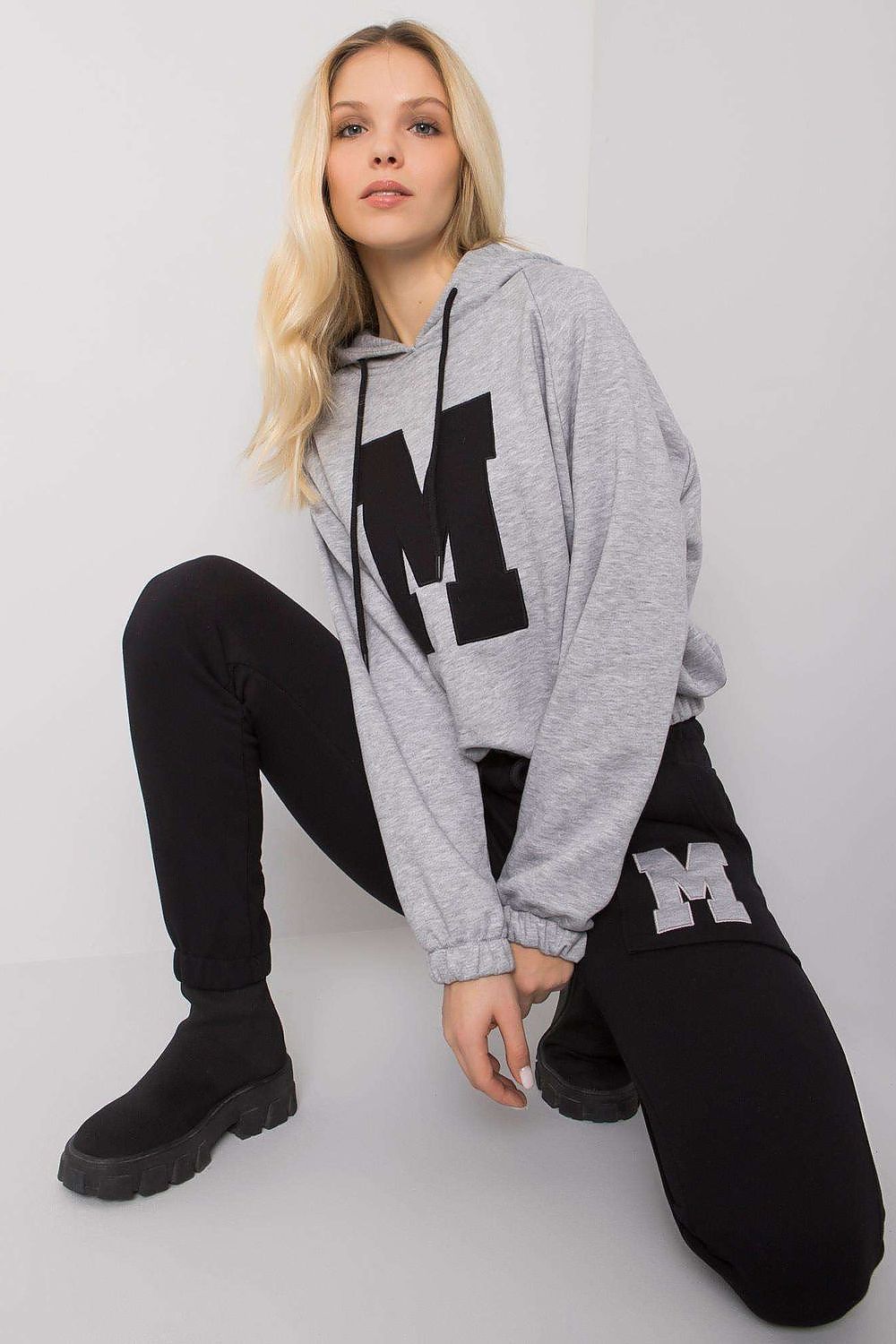 Chic women's athleisure outfit showcasing a grey hooded sweatshirt emblazoned with a bold letter 'M', styled with sleek black jogger pants, ideal for a sophisticated casual look.