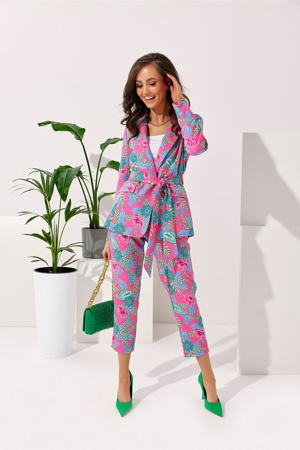 Luxurious women's Roco Fashion jacket in vibrant floral patterns, featuring decorative pockets and waist binding, styled with matching trousers and green heels, set against a minimalist background with greenery.