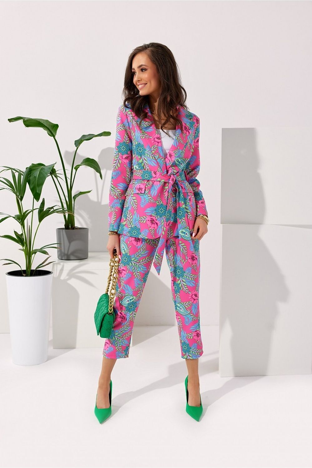 Chic woman elegantly wearing a colourful Roco fashion suit with a cinched waist and decorative details, accessorised with a green handbag and heels, surrounded by stylish interior design elements.