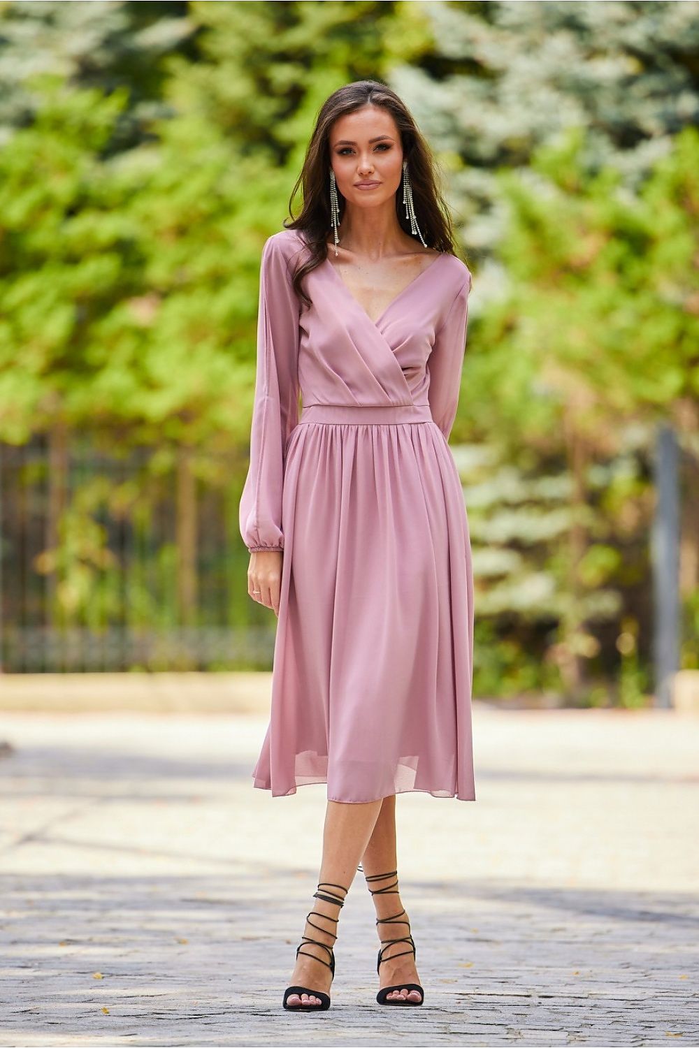 A stunning model showcasing an elegant midi cocktail dress with long buff sleeves and an exposed back in a soft pink hue, perfect for formal occasions. The dress features a flattering wrap design and is complemented by stylish strappy heels.
