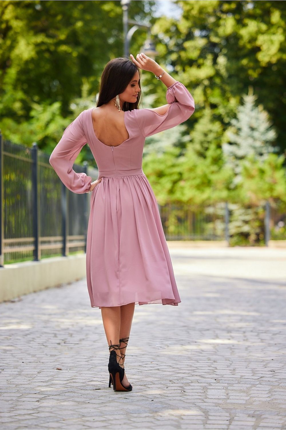 A graceful view of the back of a model in a chic midi cocktail dress with long buff sleeves and a deep V-back in pink, accentuating feminine elegance. The look is completed with fashionable strappy heels.