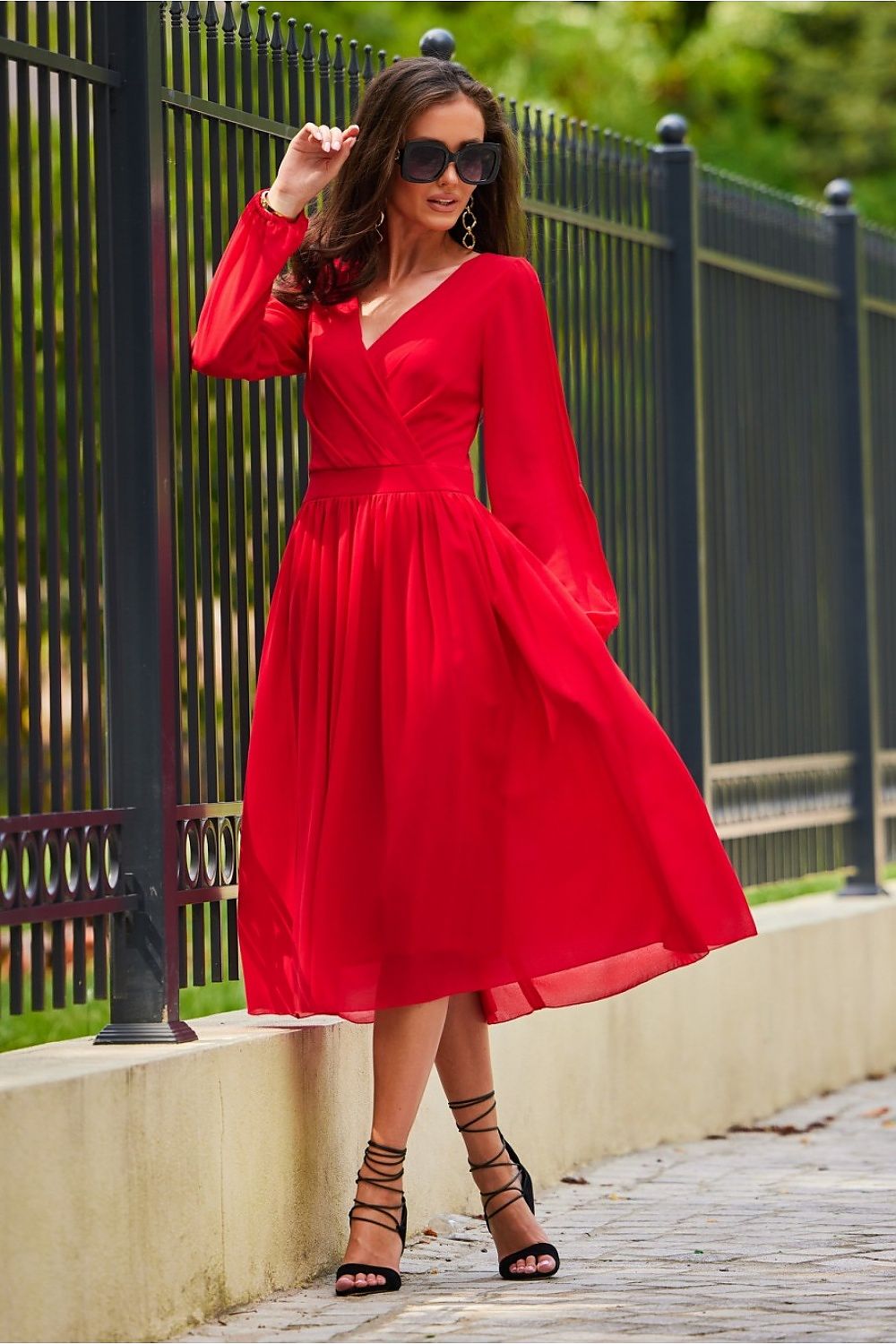 An enchanting model in a luxurious red midi cocktail dress with long sleeves and an exposed back, exuding sophistication and elegance. The dress features a wrap style complemented by delicate ankle strap heels.