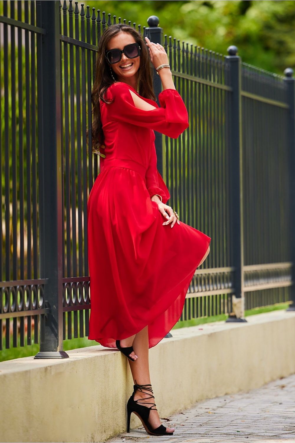 A joyful model poses in a vibrant red midi cocktail dress with elegant long sleeves and open shoulder design, paired with chic high-heeled sandals, capturing the essence of luxury and grace.