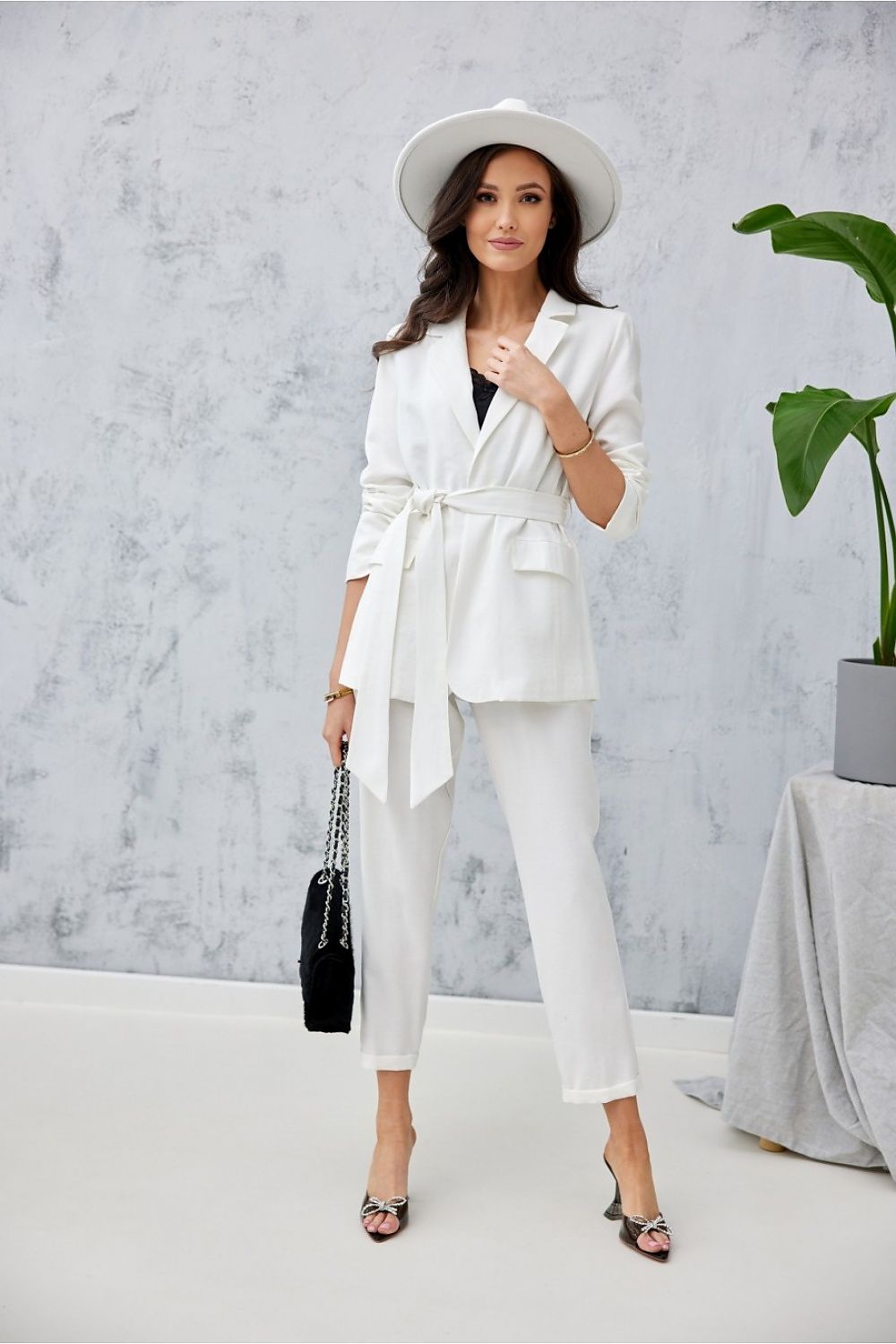 Stylish model in elegant white trousers and blazer from Roco Fashion, showcasing a sophisticated outfit complemented with a wide hat, minimalistic black bag, and fashionable heels against a modern backdrop.