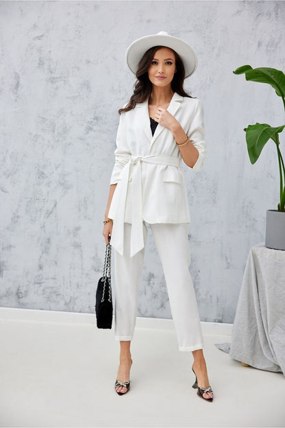 Stylish model in elegant white trousers and blazer from Roco Fashion, showcasing a sophisticated outfit complemented with a wide hat, minimalistic black bag, and fashionable heels against a modern backdrop.