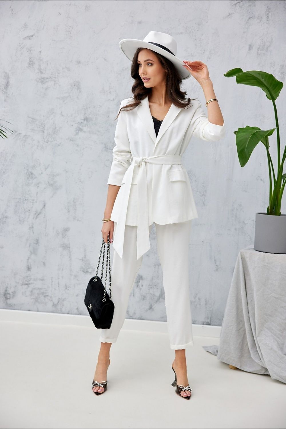 Chic woman wearing a tailored white ensemble by Roco Fashion, featuring classic trousers and a belted blazer, highlighted by a trendy hat and sleek accessories, set in a contemporary interior.