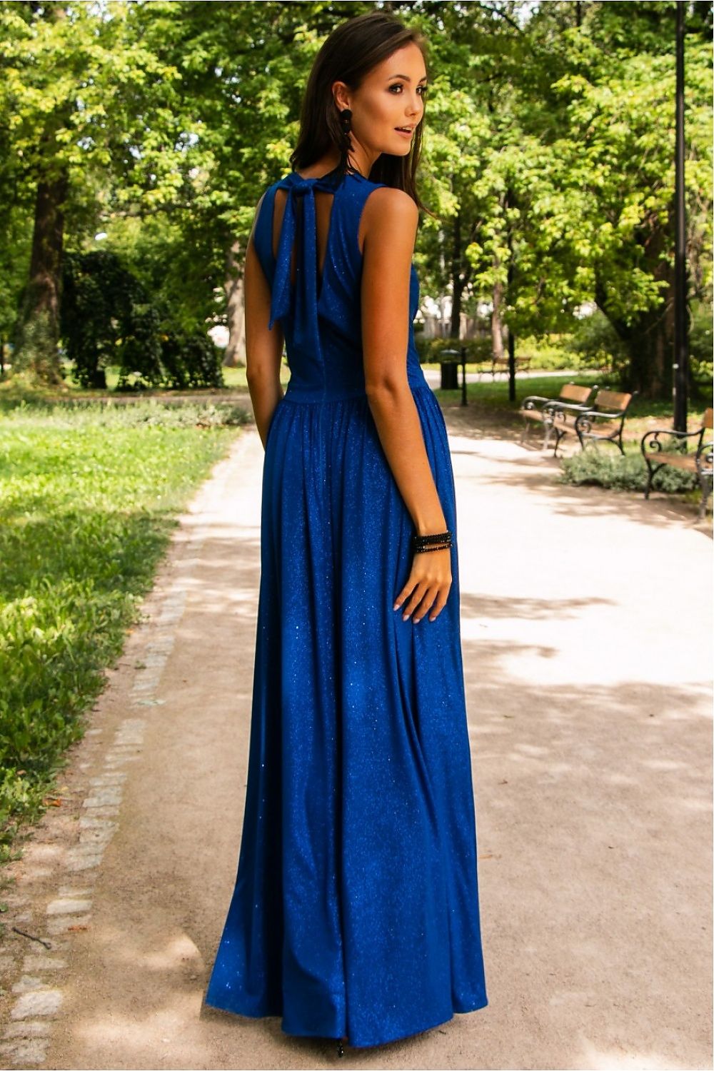 Elegant brocade maxi dress by Roco Fashion, showcasing a shimmering blue fabric with a tie back, an envelope neckline, and a stylish leg slit, perfect for evening events. The model poses gracefully in a lush park setting, highlighting the dress's flattering silhouette and luxurious details.