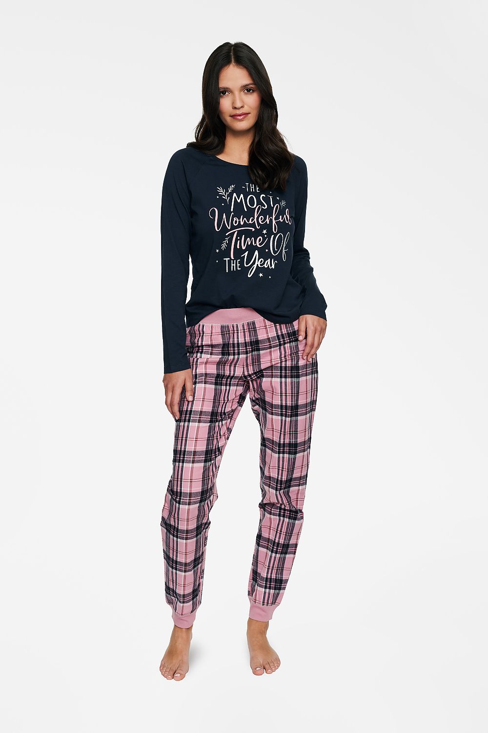 Elegant woman showcasing a luxury Christmas motif two-piece cotton pyjamas set. The long-sleeve top features a festive inscription, perfectly paired with stylish checkered pink trousers. Ideal for a comfortable, chic holiday look.
