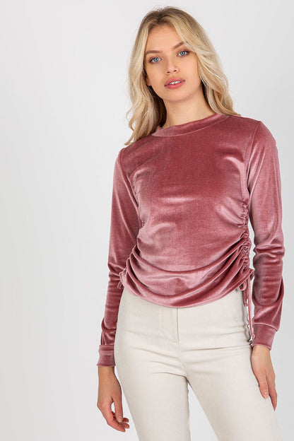 Luxurious pink velour blouse with long sleeves, featuring adjustable ties at the sides. Model showcasing a stylish look with light-coloured trousers.
