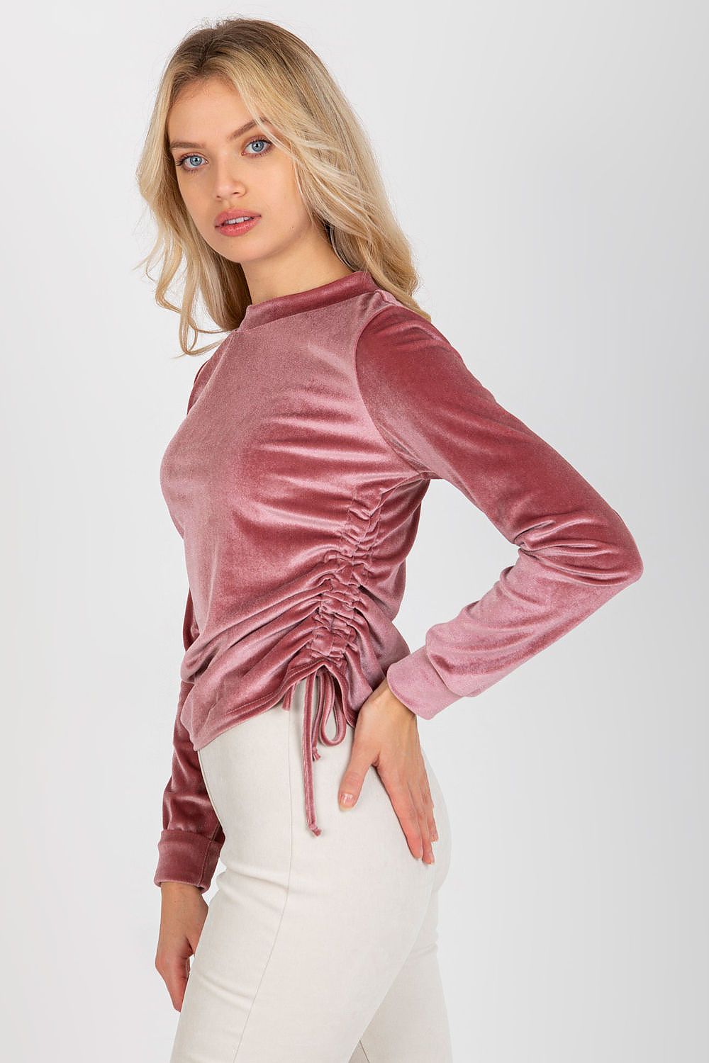 Sophisticated side profile of a model in a pink velour blouse with long sleeves and stylish ruched sides, perfectly complementing fitted trousers.