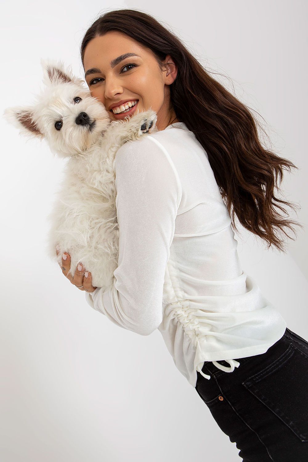 Charming model smiling while holding a fluffy white dog, wearing a white luxurious velour blouse with long sleeves and side ties, exuding warmth and elegance.