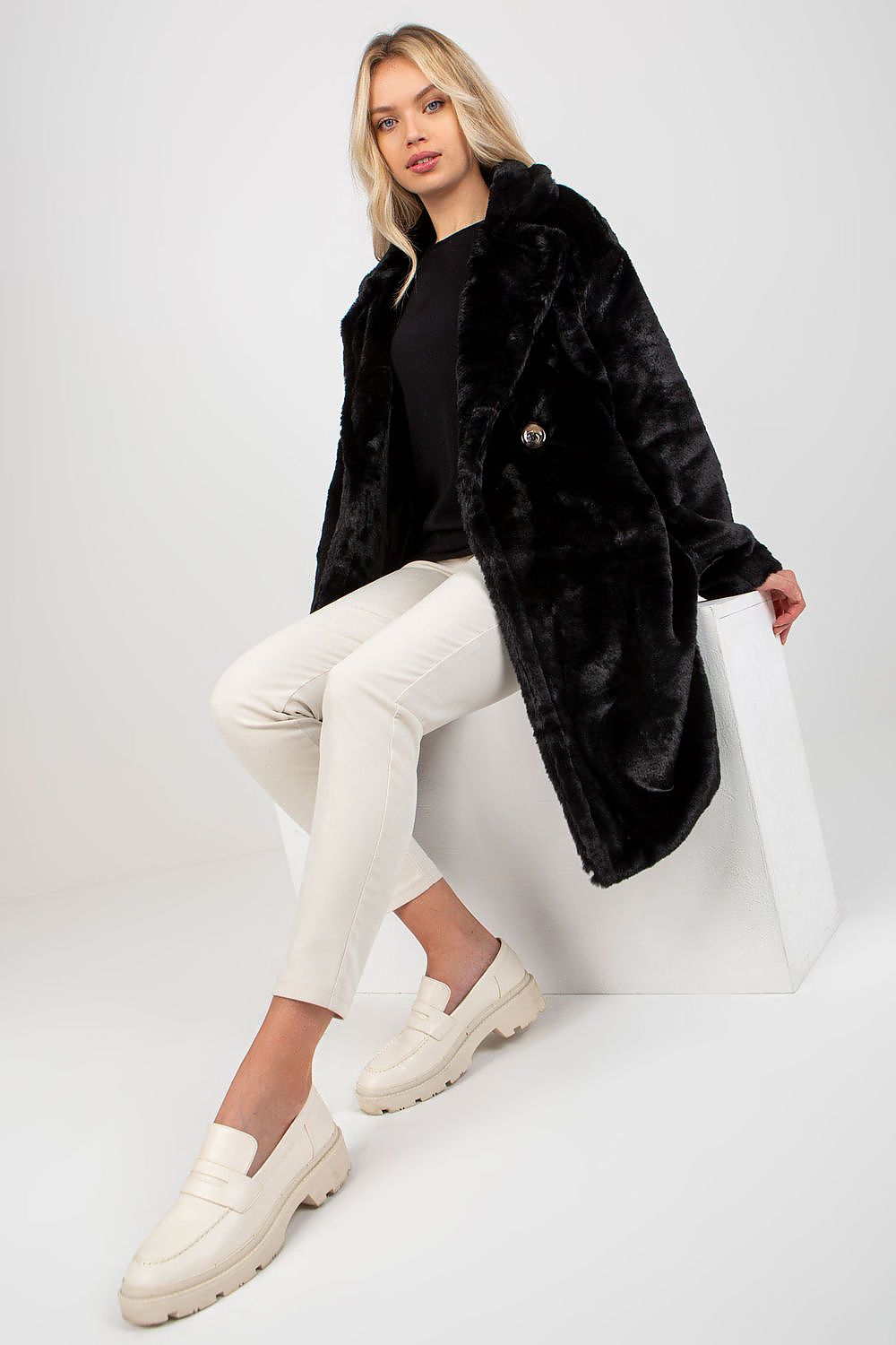 Stylish model in a black fur coat by Och Bella, sitting confidently while wearing elegant white trousers and a black top, emphasizing the coat's plush material.