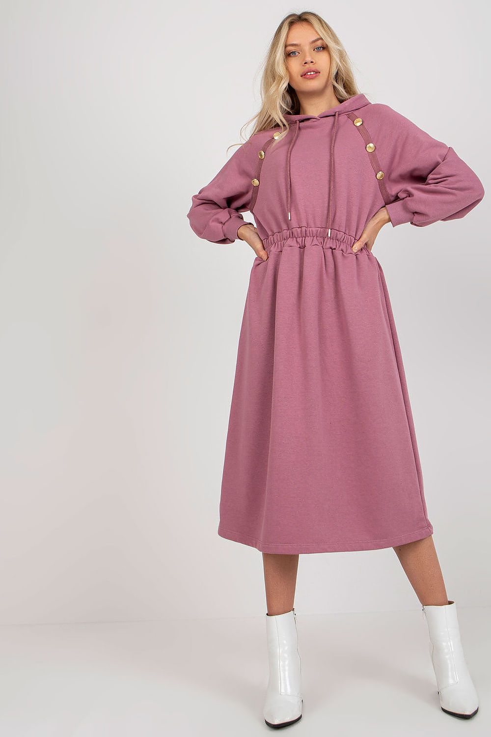 Fashionable woman presenting a charming pink cotton hoodie dress with decorative buttons and a cinched waist, paired with sleek white ankle boots, perfect for a trendy, casual ensemble.