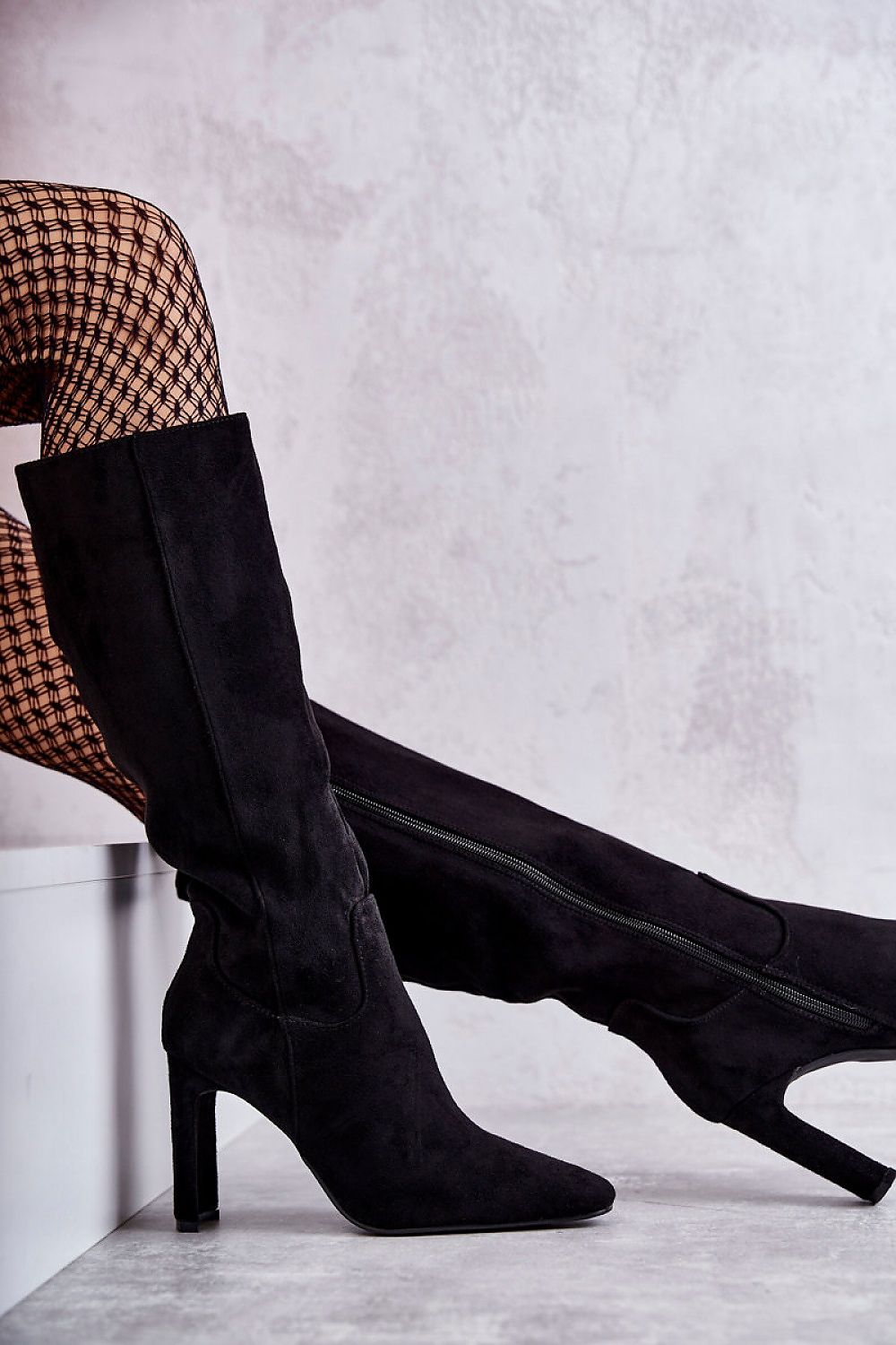 Sophisticated black eco-suede knee-high boots showcasing a side zip detail and pointed toe, accented by fishnet stockings against a sophisticated backdrop.