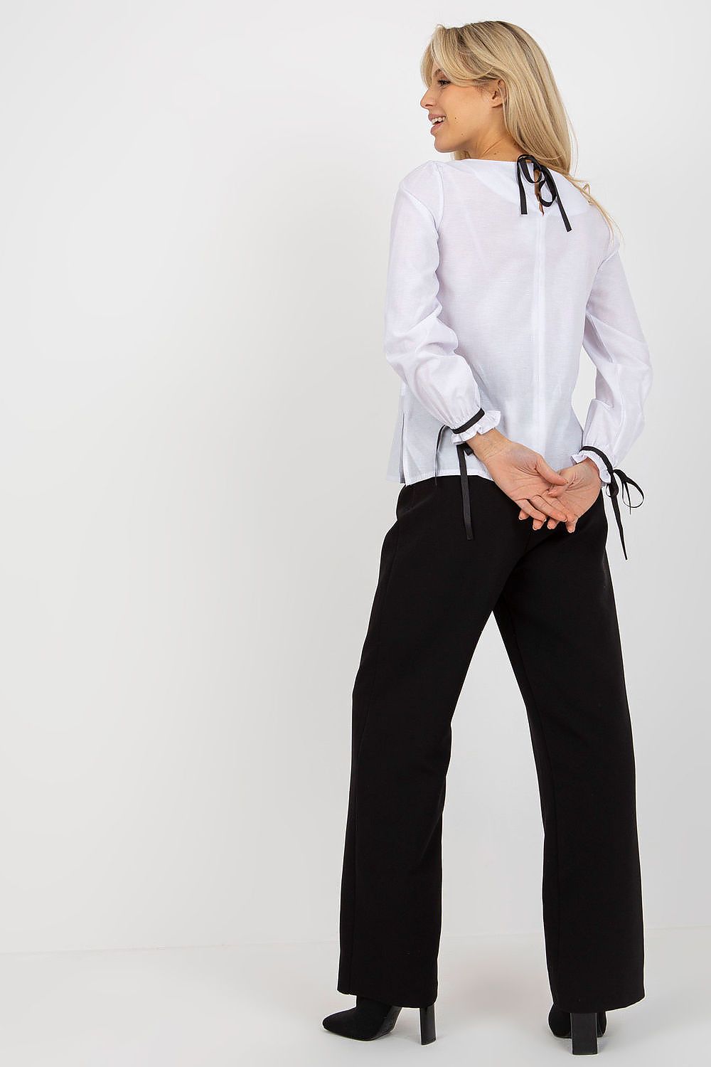 Back view of a sophisticated white luxury cotton blouse with lace detailing, featuring elegant tying closure, paired with trendy black trousers.