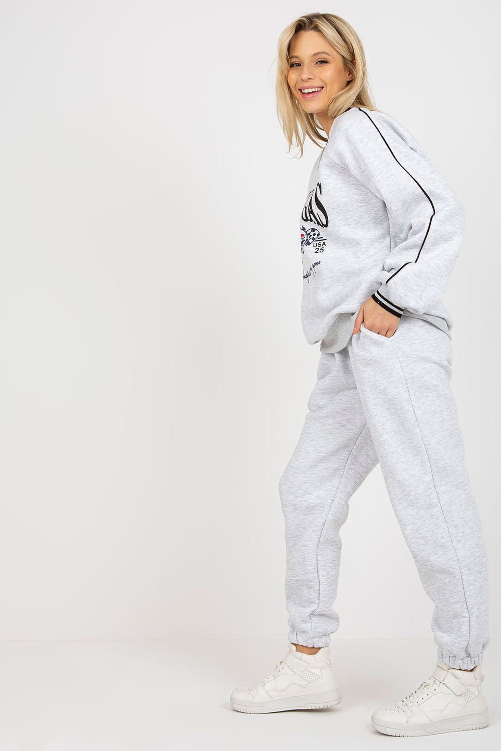 Stylish model showcasing a grey sweatshirt and matching pants set, smiling radiantly while standing confidently in chic white footwear, embodying modern elegance.