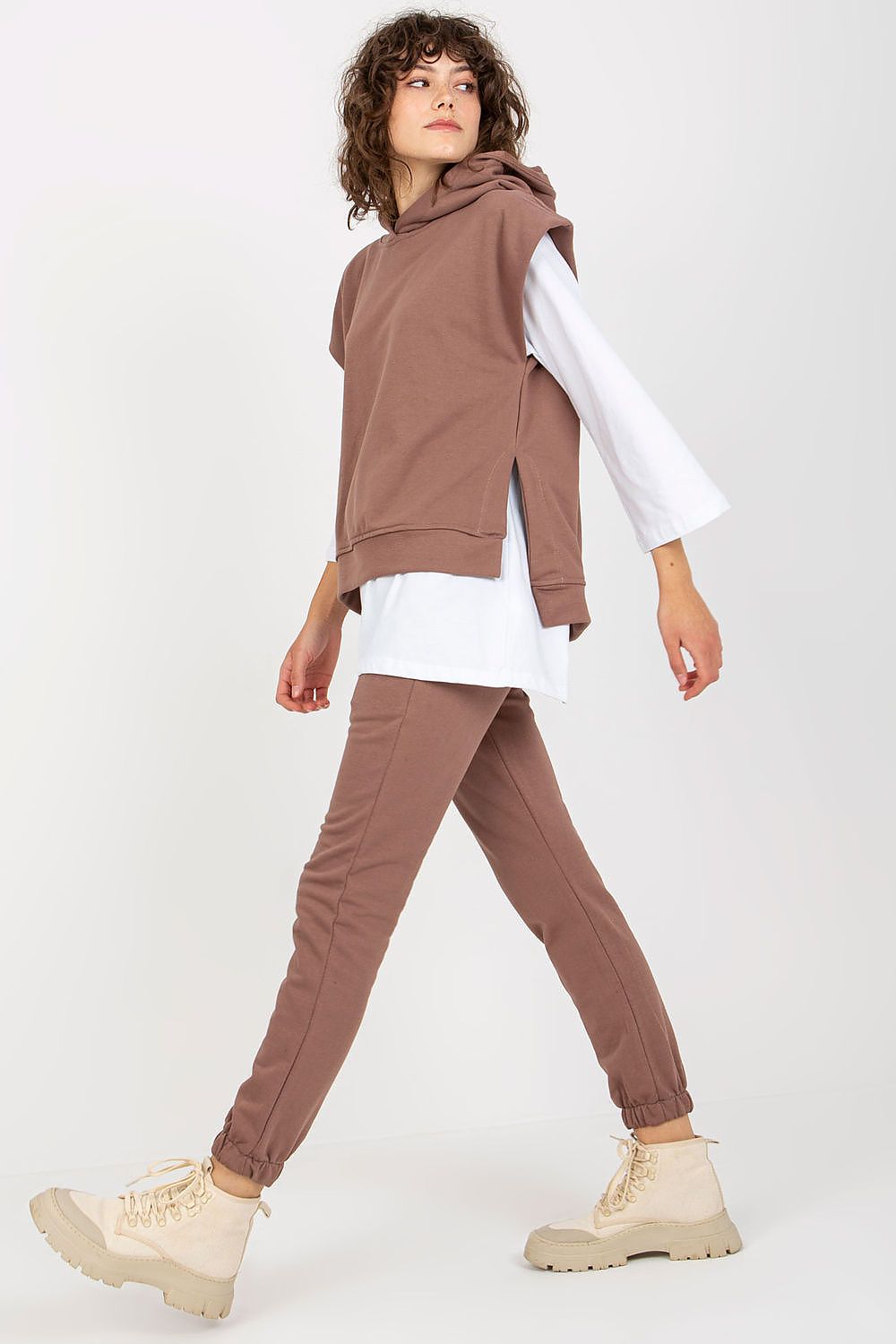 Stylish model showcasing a fashionable brown sweatsuit set, with a sleeveless hoodie over a white blouse, paired with tailored pants, exuding a sense of modern elegance.