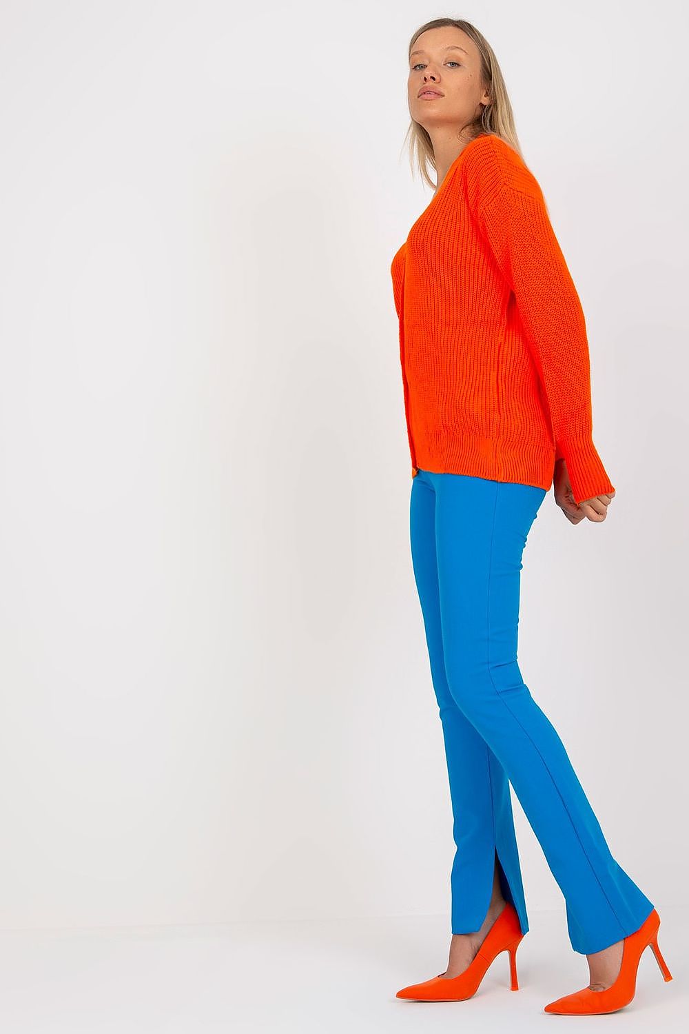 Side view of a stylish Rue Paris Women's acrylic cardigan in bright orange, complemented by blue trousers and high-heeled shoes, highlighting modern fashion trends.