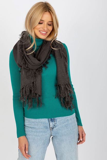A stylish woman wearing a luxurious 100% acrylic shawl scarf in dark grey with elegant tassels, complemented by a vibrant teal ribbed top and light-wash jeans, showcasing an upscale fashion statement.