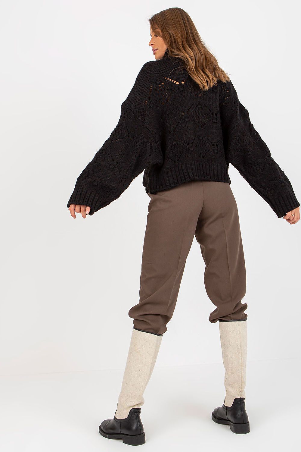 Back view of a model wearing a sophisticated black openwork turtleneck sweater with wide sleeves, complemented by tailored trousers and stylish boots.