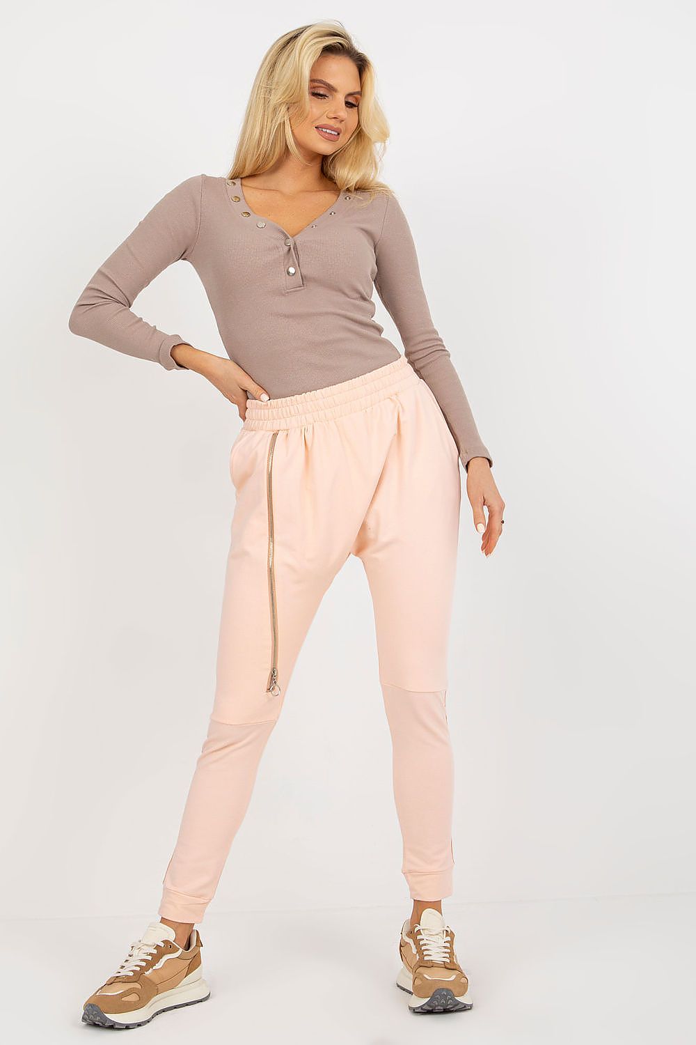 Elegant model in high waist tracksuit trousers with decorative zipper and side pockets, styled with a chic beige top and trendy sneakers, showcasing stylish comfort.